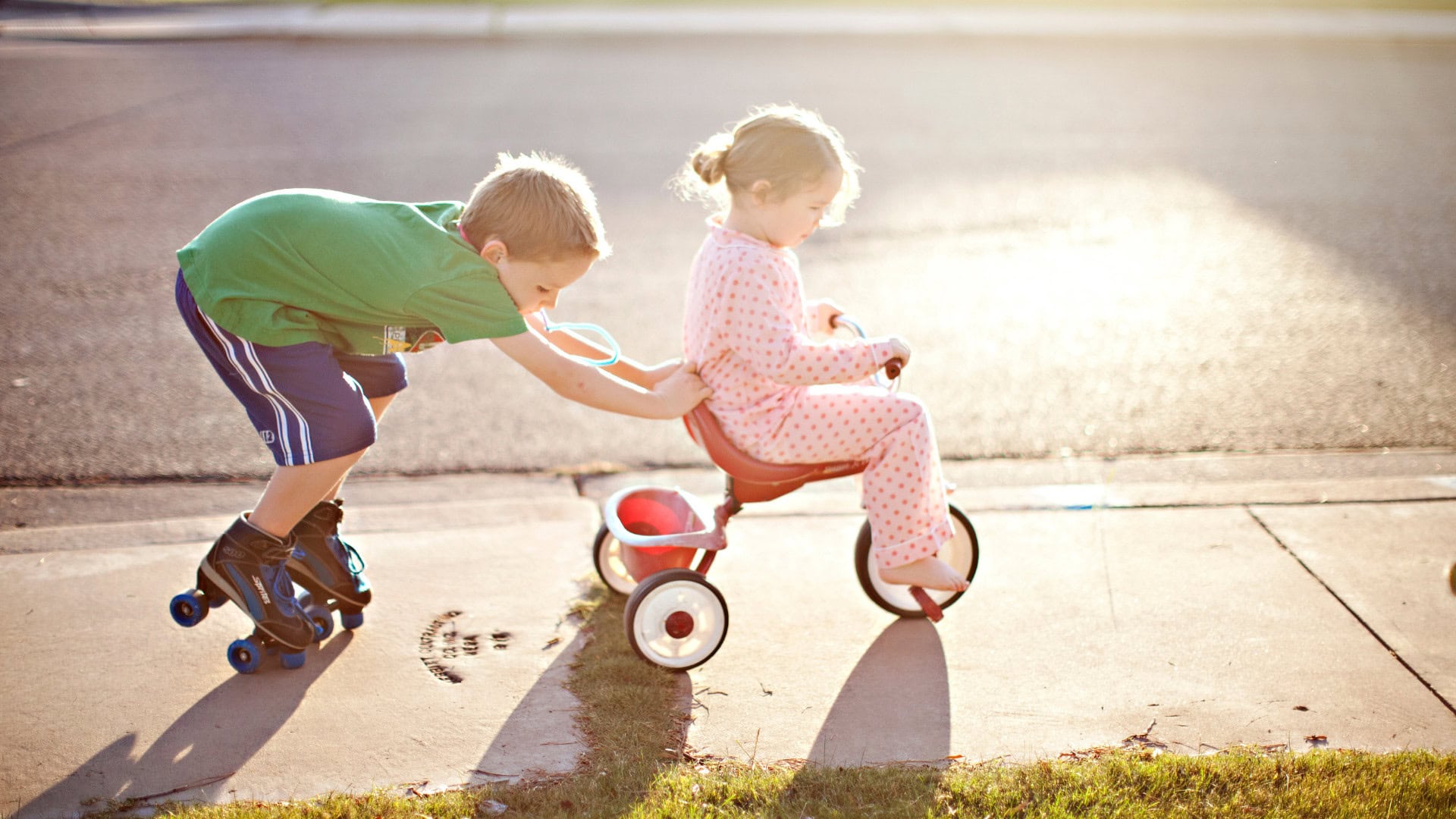 Young boy pushes girl on a tricycle on the sidewalk during daytime.