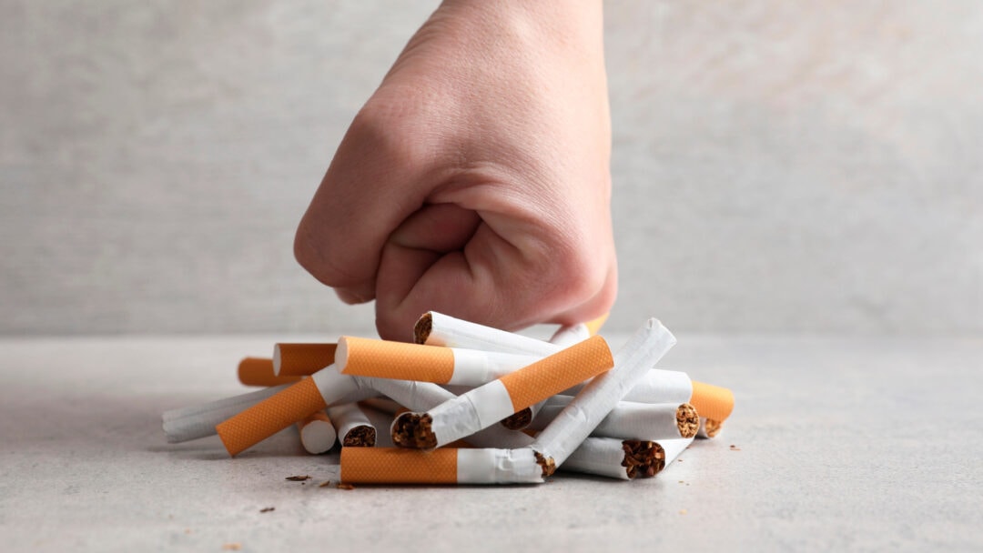 Person's hand crushing a pile of cigarette butts and cigarette butts on the ground.